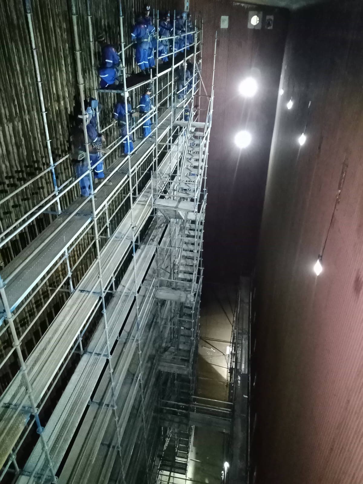 Equipe com EPI trabalhando em andaimes de vários níveis no interior de uma grande estrutura confinada iluminada